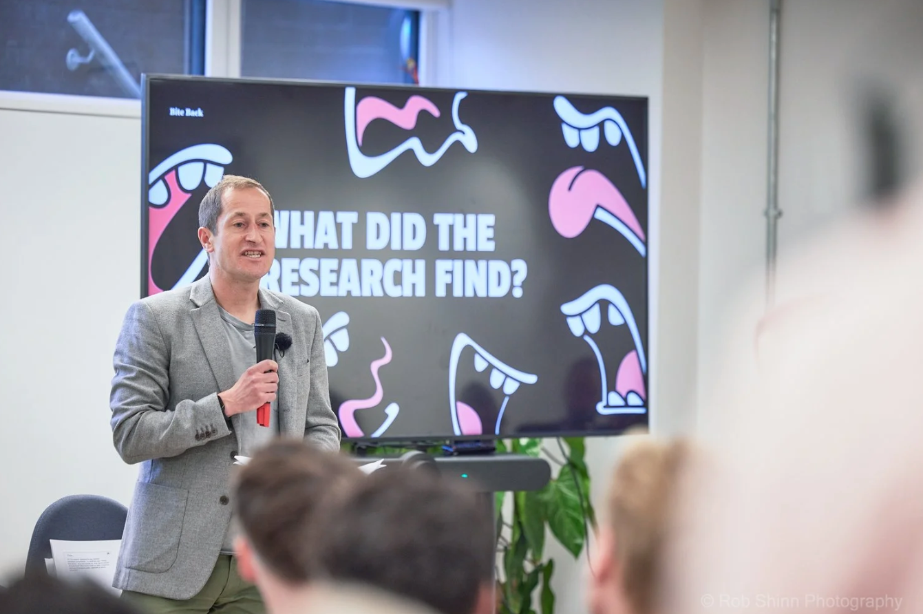 Professional speaker presenting at a business conference with digital screens displaying research findings in the background.