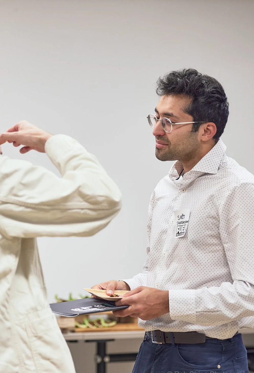 A person in a white polka dot shirt and glasses gestures while holding papers in a classroom or studio setting.