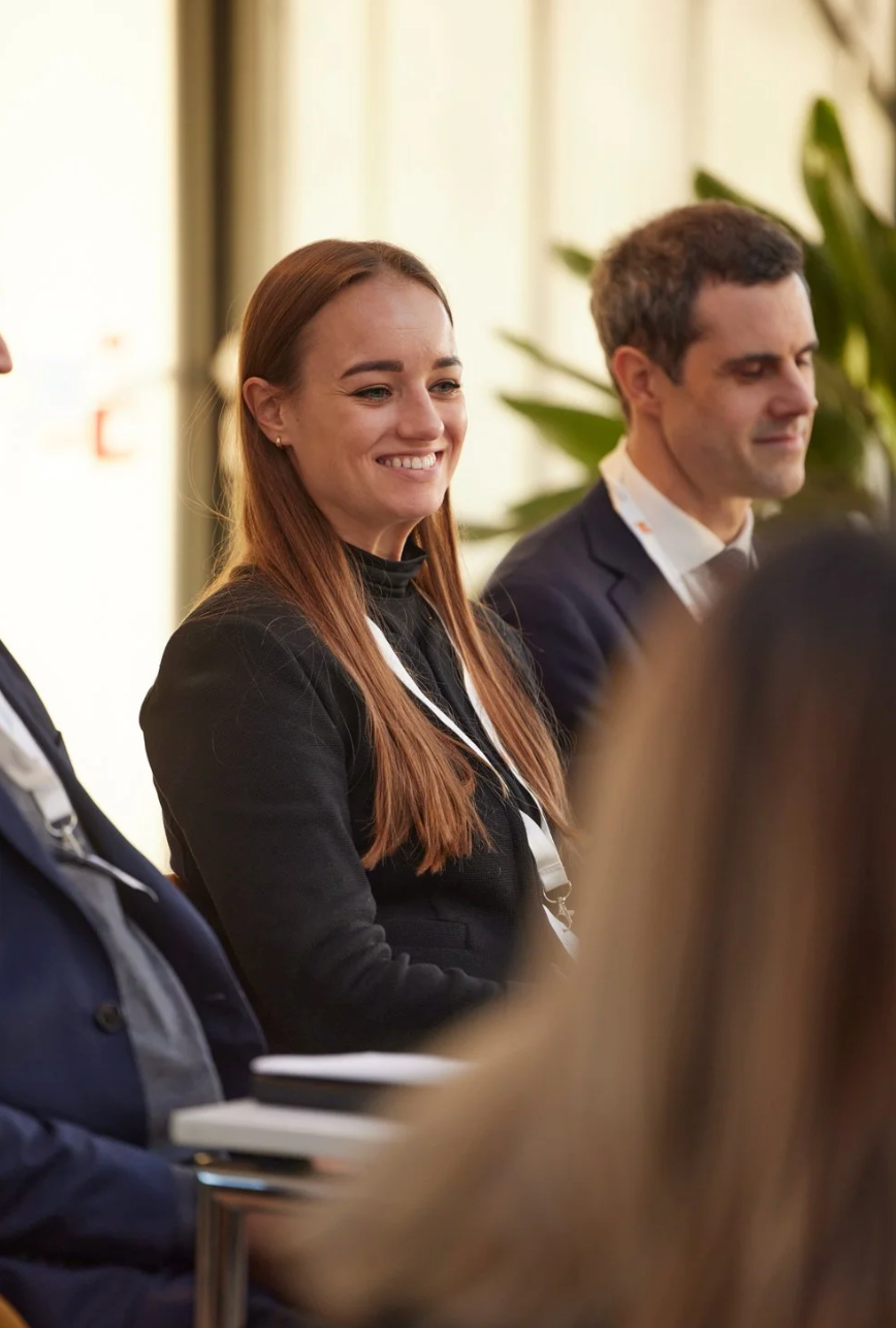 Business professionals in formal attire smile and engage during a meeting in a bright conference room setting.