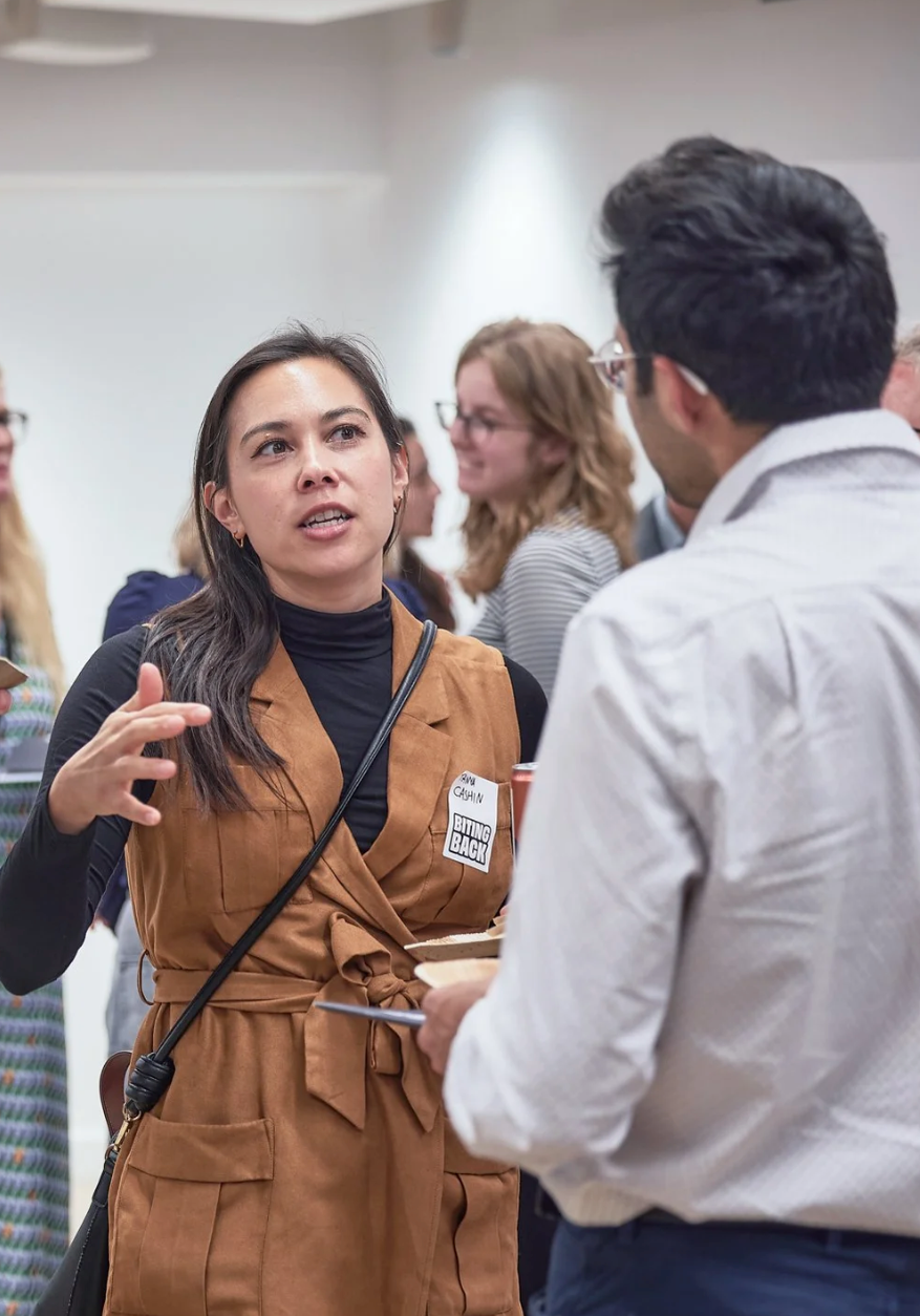 People engage in animated conversation at a professional networking event, with someone wearing a brown coat gesturing while speaking.