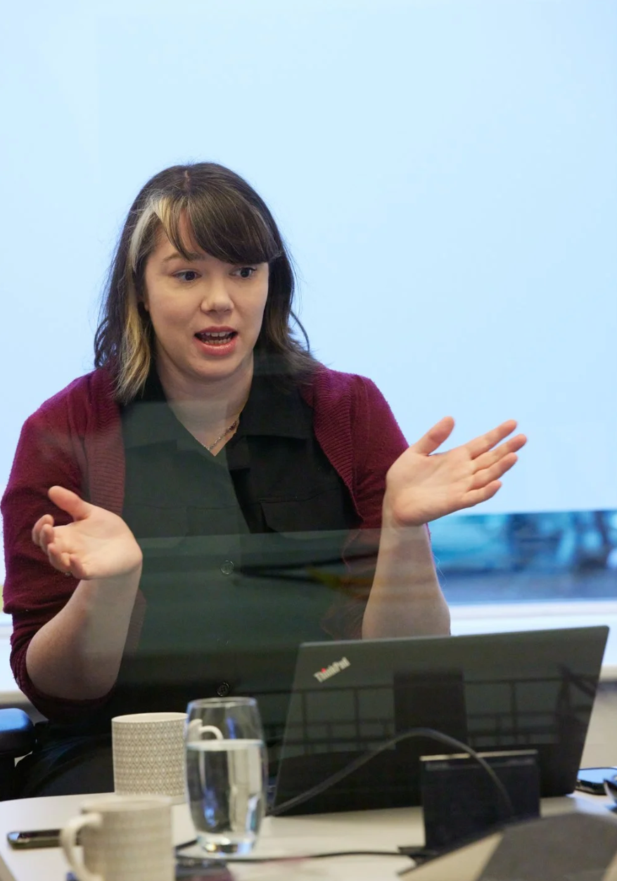 A person in a burgundy sweater gestures animatedly while sitting at a desk with a laptop during a meeting.