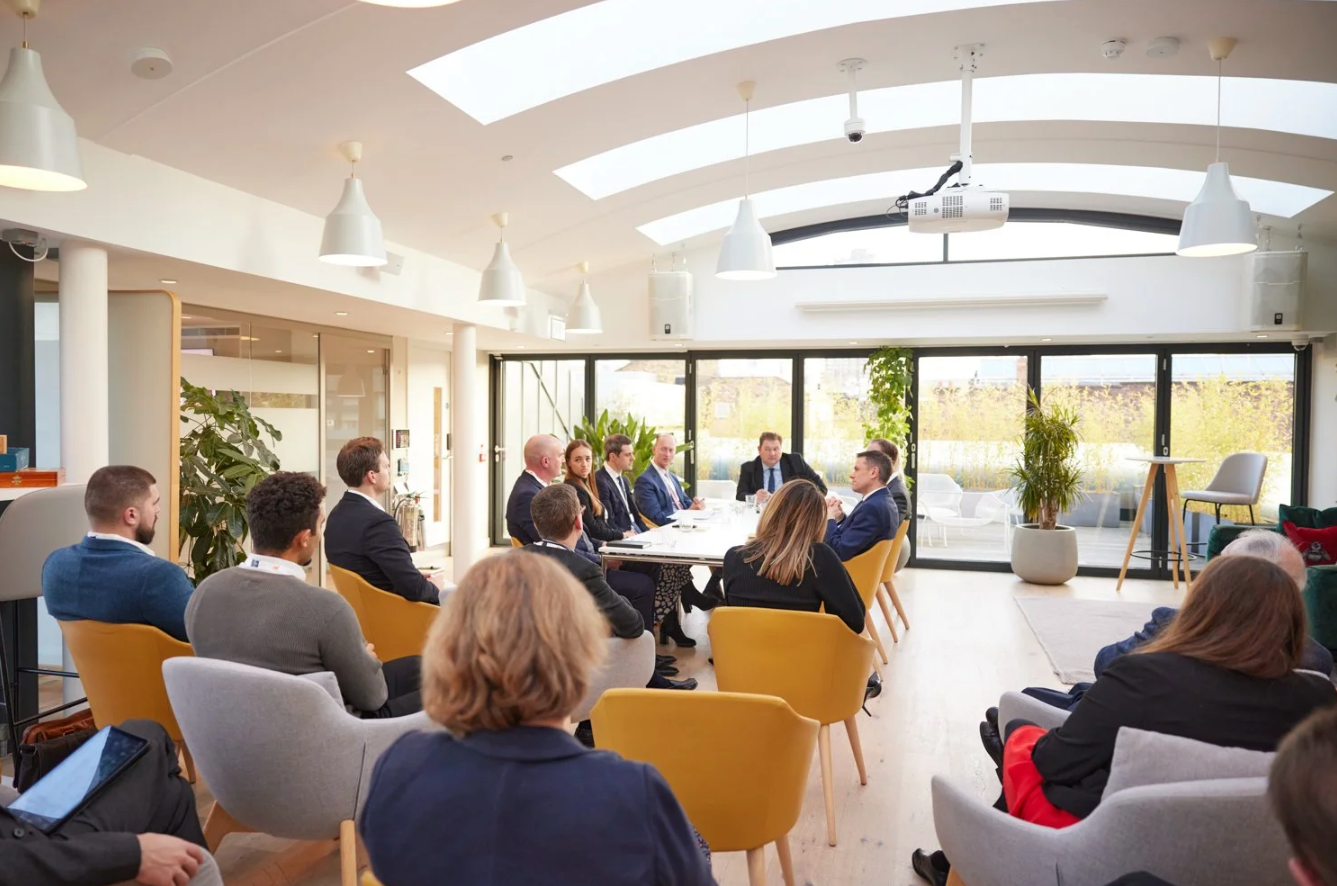 Audience members seated in yellow and gray chairs listen attentively during a bright indoor presentation space.