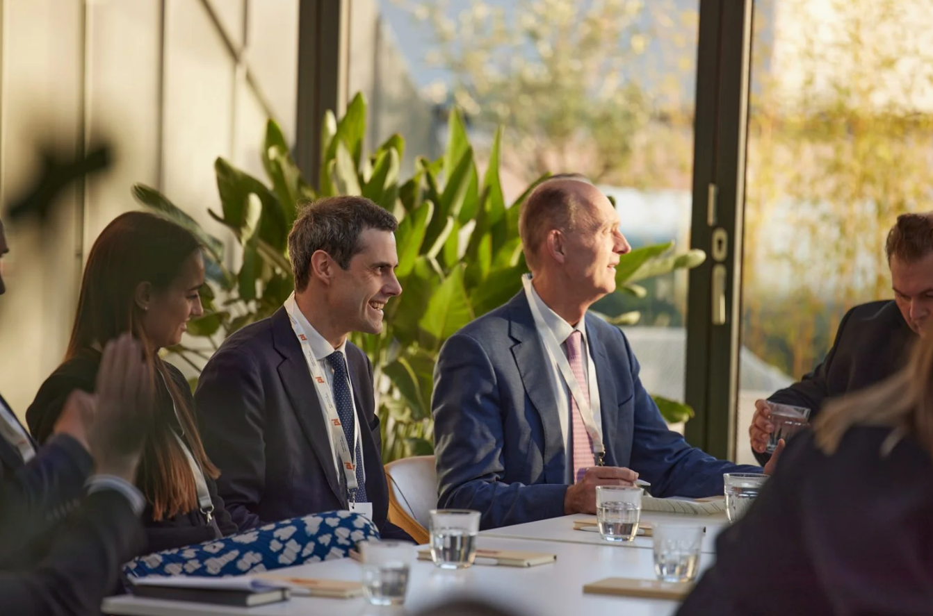 Business professionals seated at tables during a corporate meeting or conference in a bright modern space.