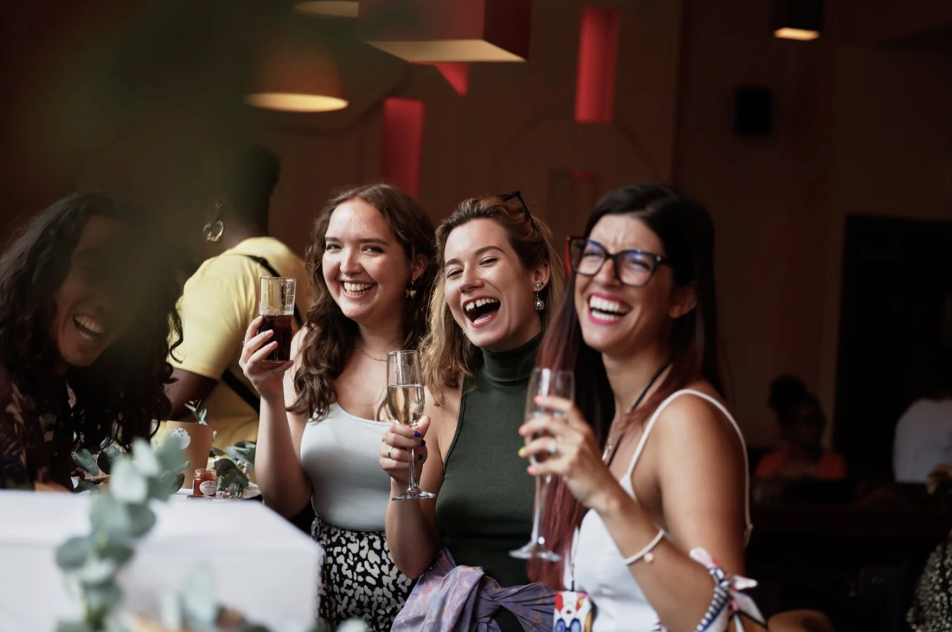 A group of wedding guests laugh together at a reception table while enjoying drinks and conversation.