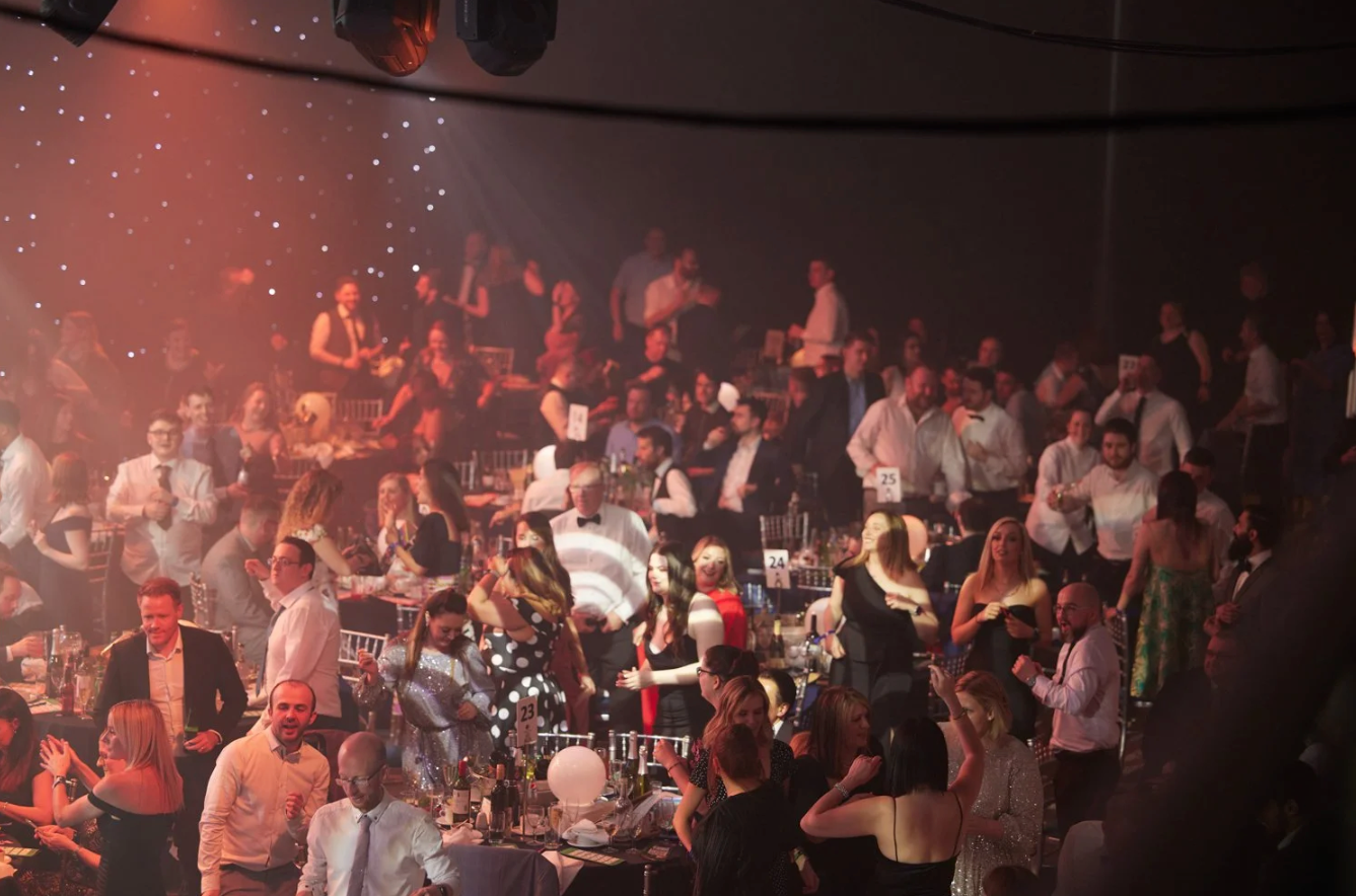 A crowded concert venue with red lighting shows an energetic audience enjoying live music from the stands.
