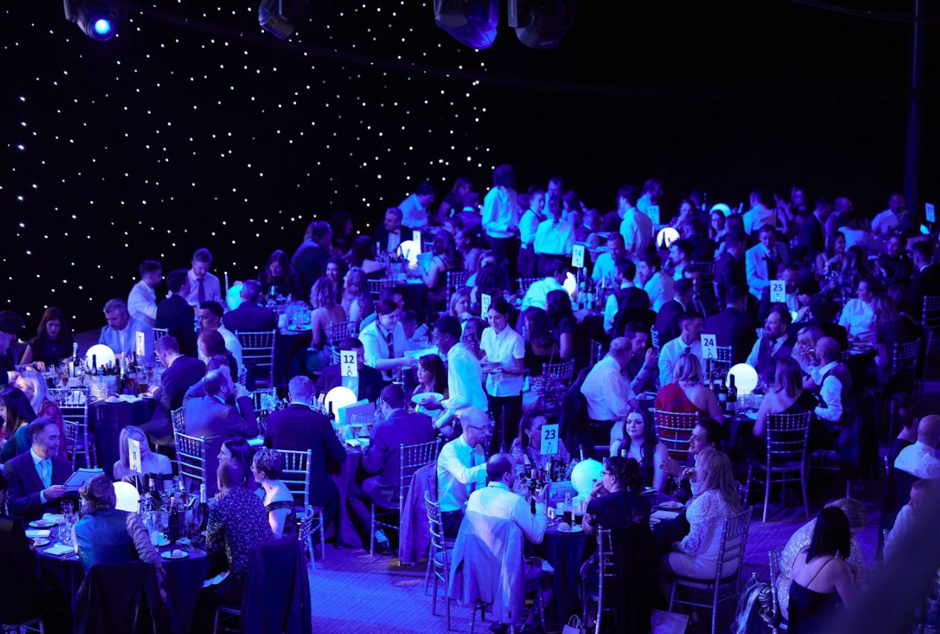 Concert audience sits in dimly lit venue with blue stage lighting creating an atmospheric ambiance during performance.