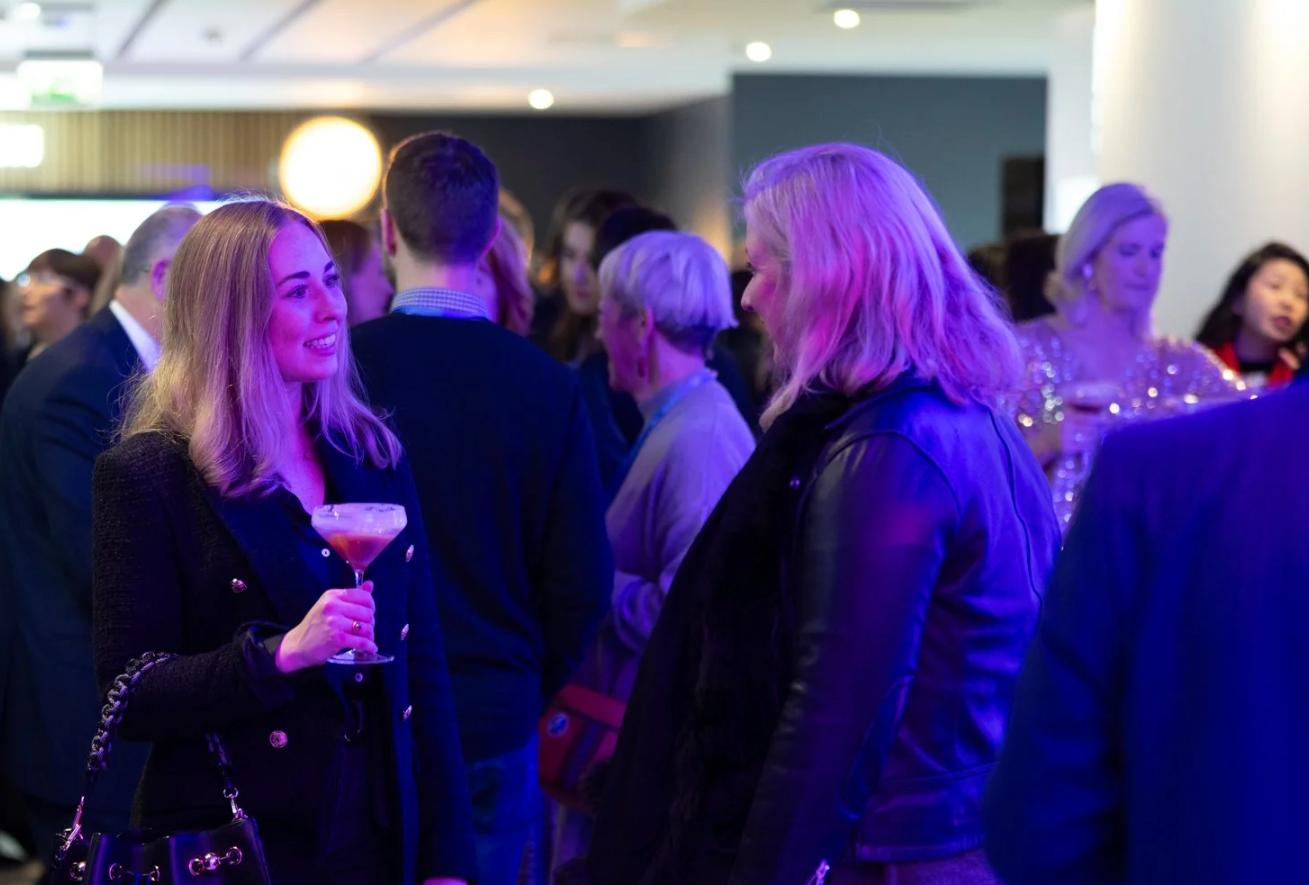 People enjoying a nightclub atmosphere with purple lighting and cocktails at a crowded social gathering.