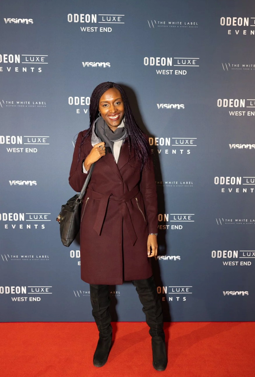 A person in a burgundy suit and black shoes poses on a red carpet against a backdrop with Odeon branding.