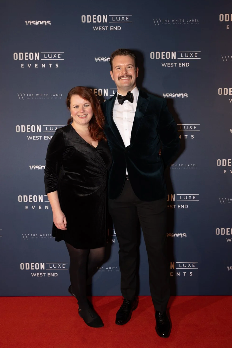 Two people pose together on a red carpet at an Odeon Luxe event with a dark branded backdrop.