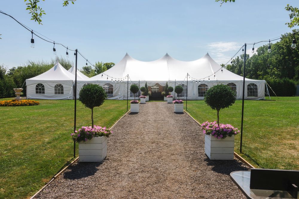 A gravel pathway leads to elegant white wedding tents with decorative planters and string lights in an outdoor garden setting.