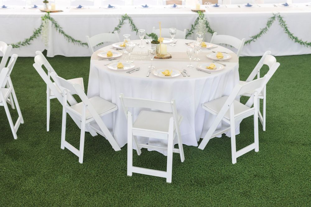 Outdoor wedding reception setup with white round tables, chairs and tent decorations on a lush green lawn.