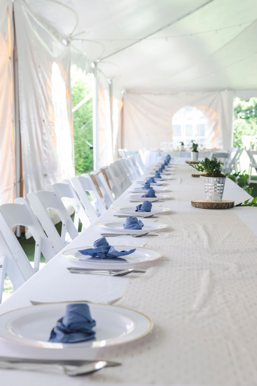 Long banquet table with elegant place settings featuring white tablecloths and navy blue napkins under a tent.
