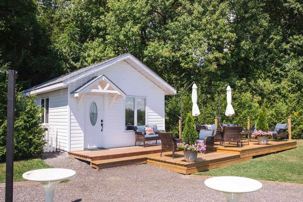A panoramic view of a white house with a deck surrounded by lush green trees and landscaping.