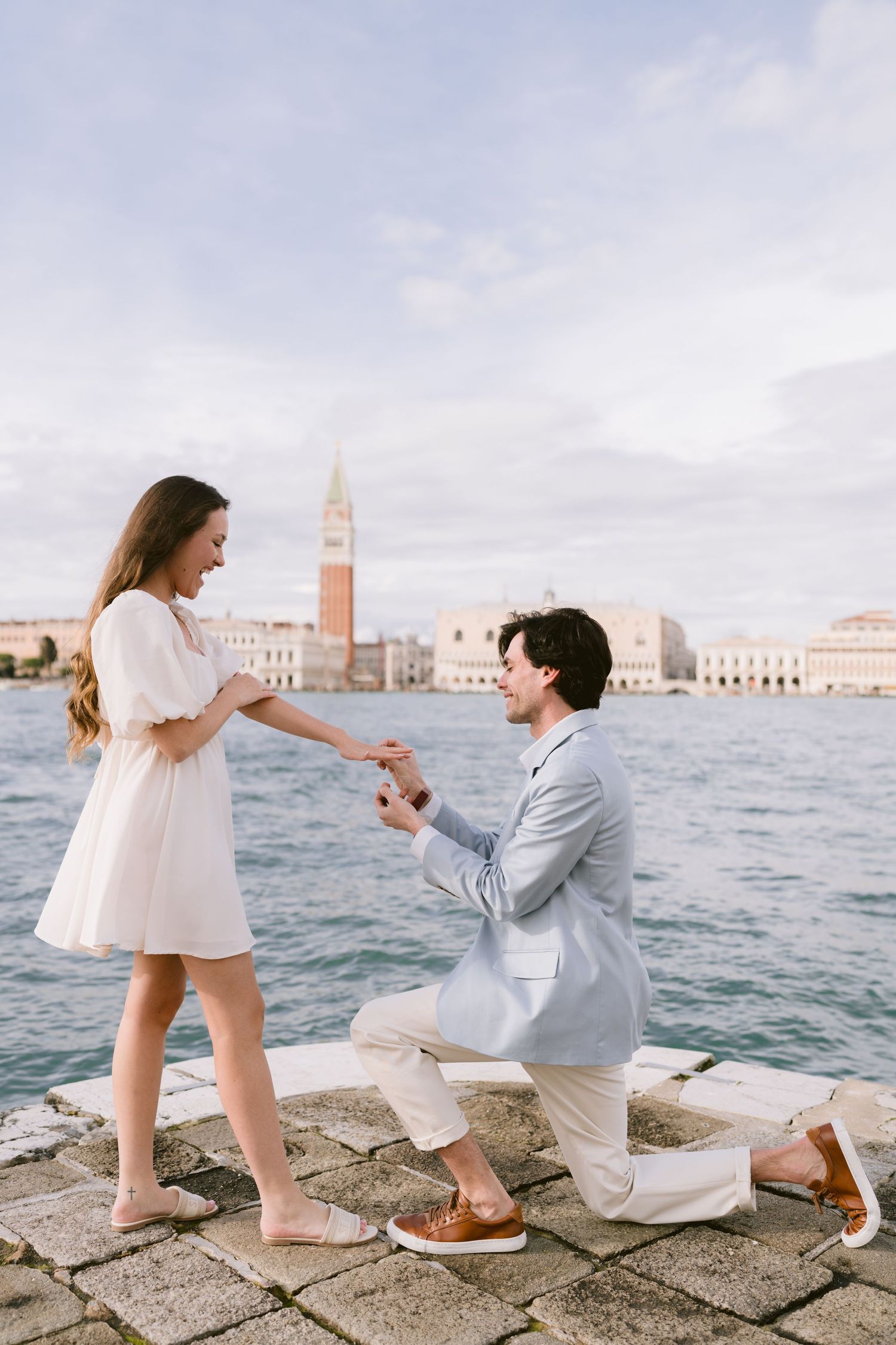 A romantic proposal sequence unfolds on a stone pier overlooking the Venice lagoon and cityscape. Surprise proposal photographer Alina Indi.