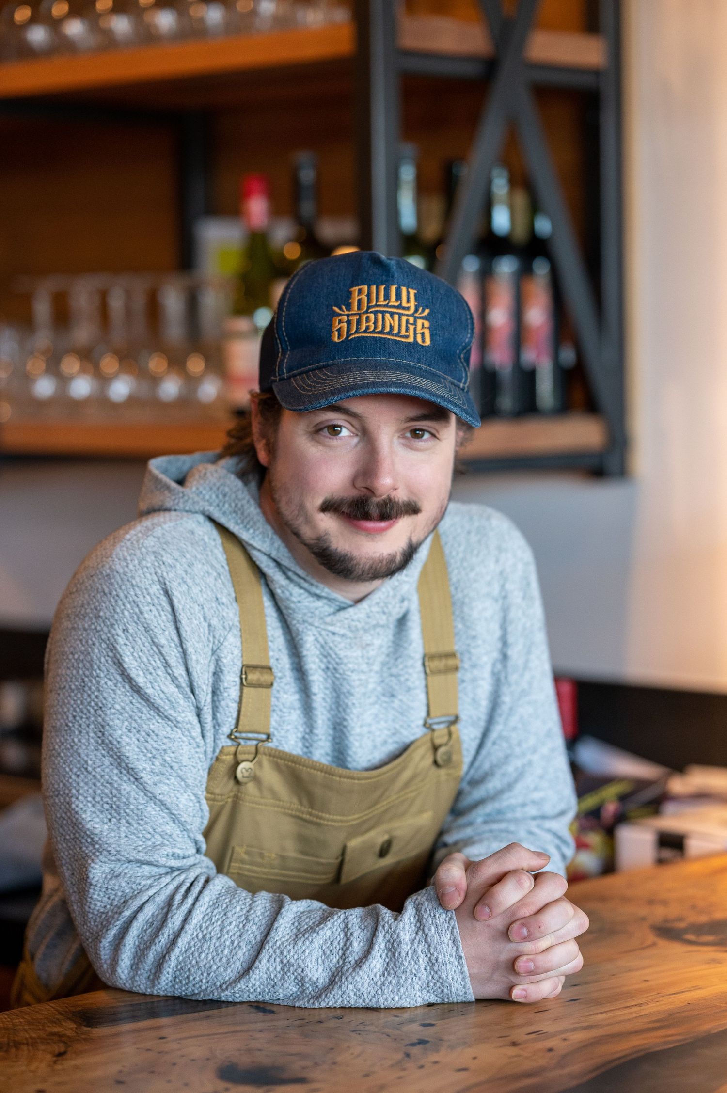 Staff member in khaki overalls and baseball cap rests on wooden bar counter.