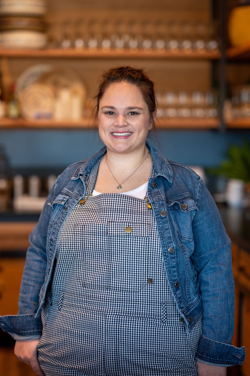 Person wearing a gingham apron and denim jacket posing in a restaurant setting.