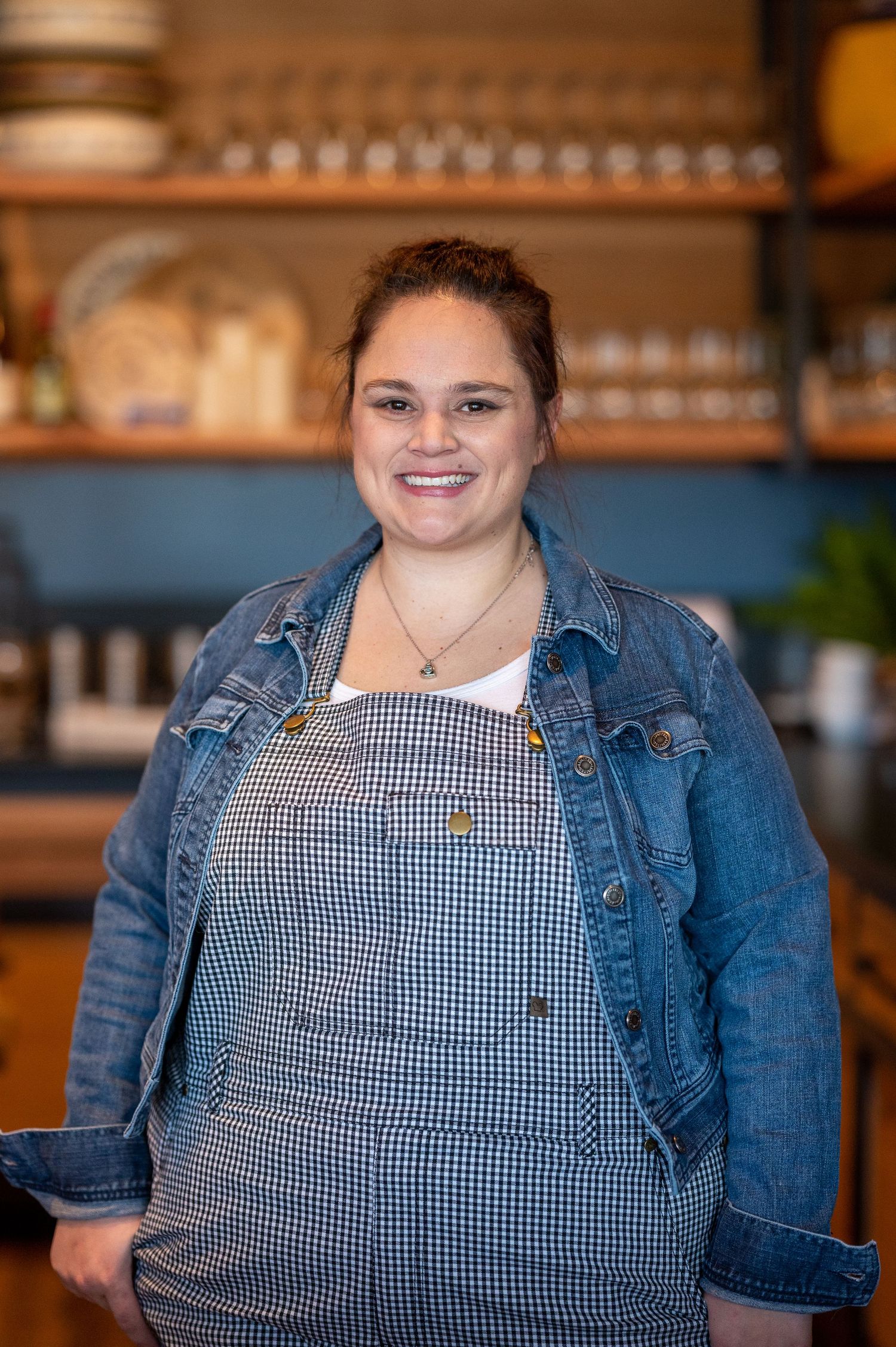 Person wearing a gingham apron and denim jacket posing in a restaurant setting.