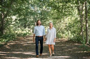 A couple walks hand in hand down a tree-lined dirt path on a sunny afternoon.
