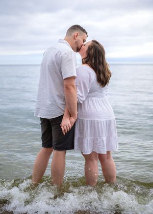 A couple in white and black outfits stands together in the ocean waves at sunset, creating a romantic seaside moment.