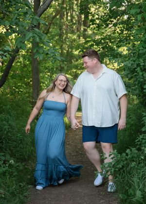 A couple in blue and white outfits walk hand in hand along a wooded trail on a sunny summer day.