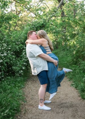 A couple embraces and lifts each other on a dirt path surrounded by lush green foliage on a sunny day.
