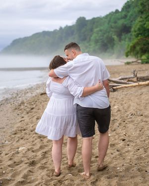 A couple embraces while walking barefoot on a misty beach with forested cliffs in the background.