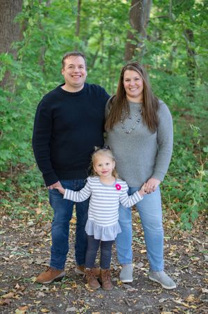 A family poses together outdoors in a wooded area with fallen autumn leaves on the ground.