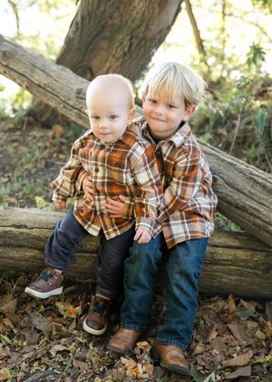 A child and sibling sit together on fallen logs surrounded by autumn leaves in a natural outdoor setting.