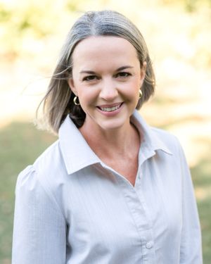 A person in a light gray sweater and white collared shirt smiles warmly against a soft blurred background.