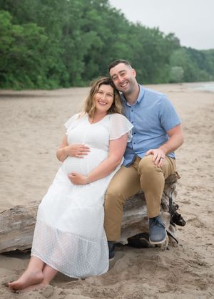 A couple sits together on driftwood at a sandy beach during an overcast day.