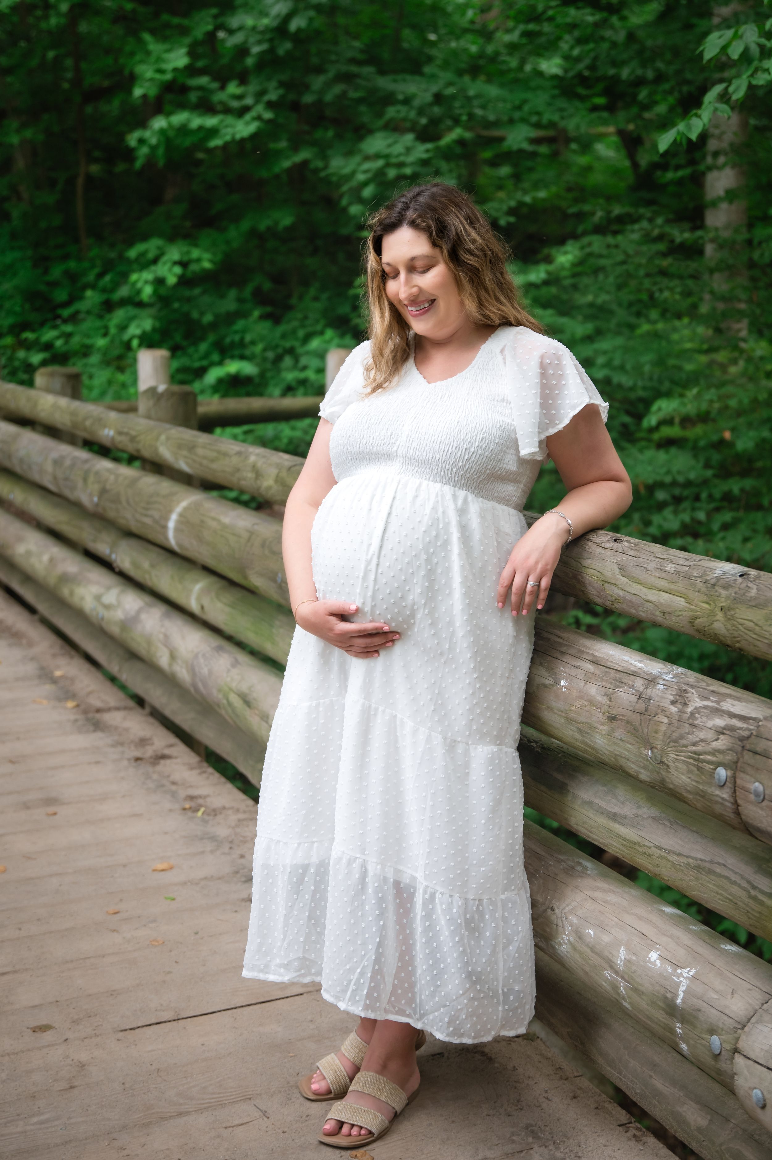 A pregnant woman in a flowing white dress stands on a wooden bridge surrounded by lush green forest.