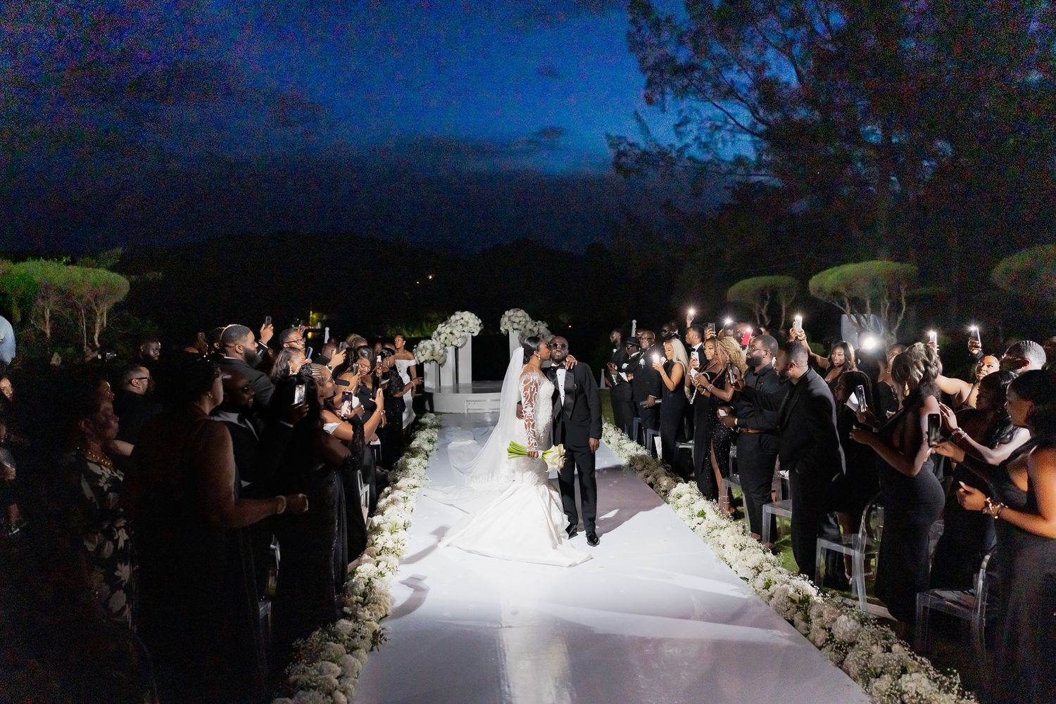 Wedding guests gather outdoors at dusk for a nighttime celebration with twinkling lights and decorative elements.