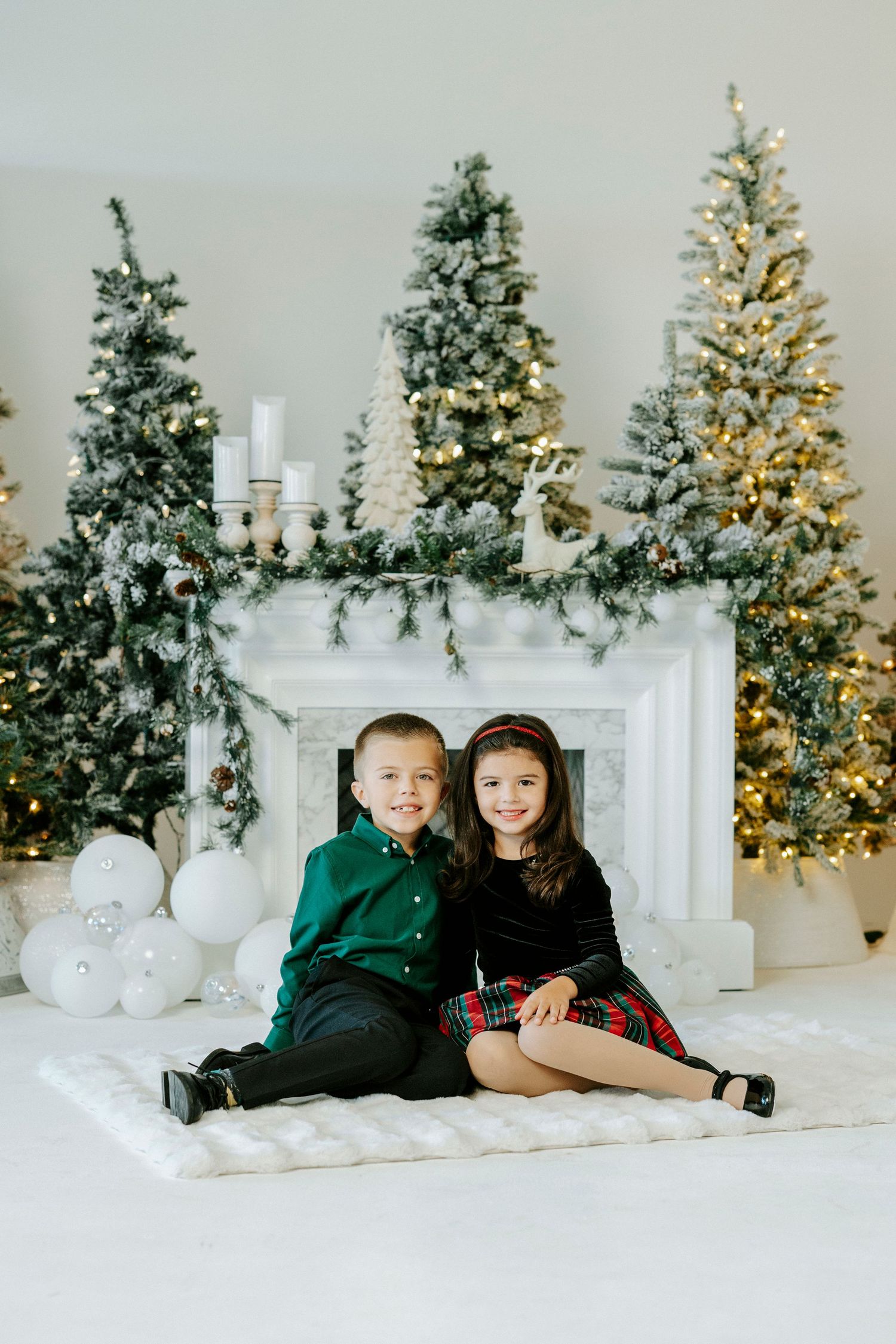 Children in holiday attire pose together on a white floor with decorated Christmas trees and a fireplace in the background.