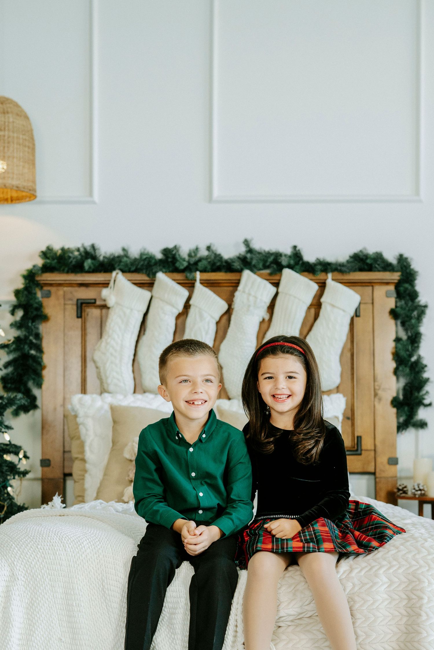 Two children sit together on a white bed with holiday decor and greenery garland in the background.