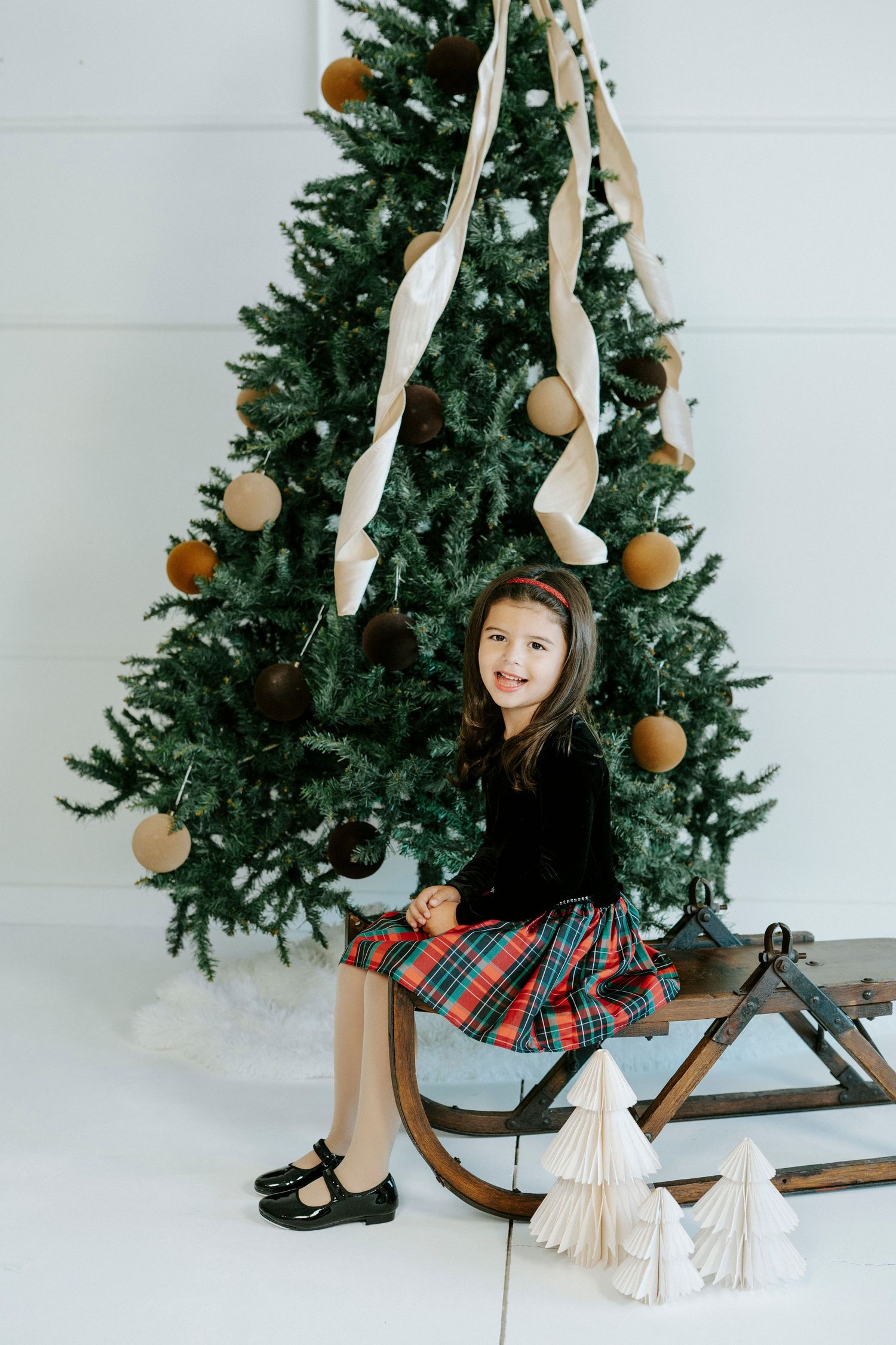 A child sits on a wooden sled next to a decorated Christmas tree in a white room.