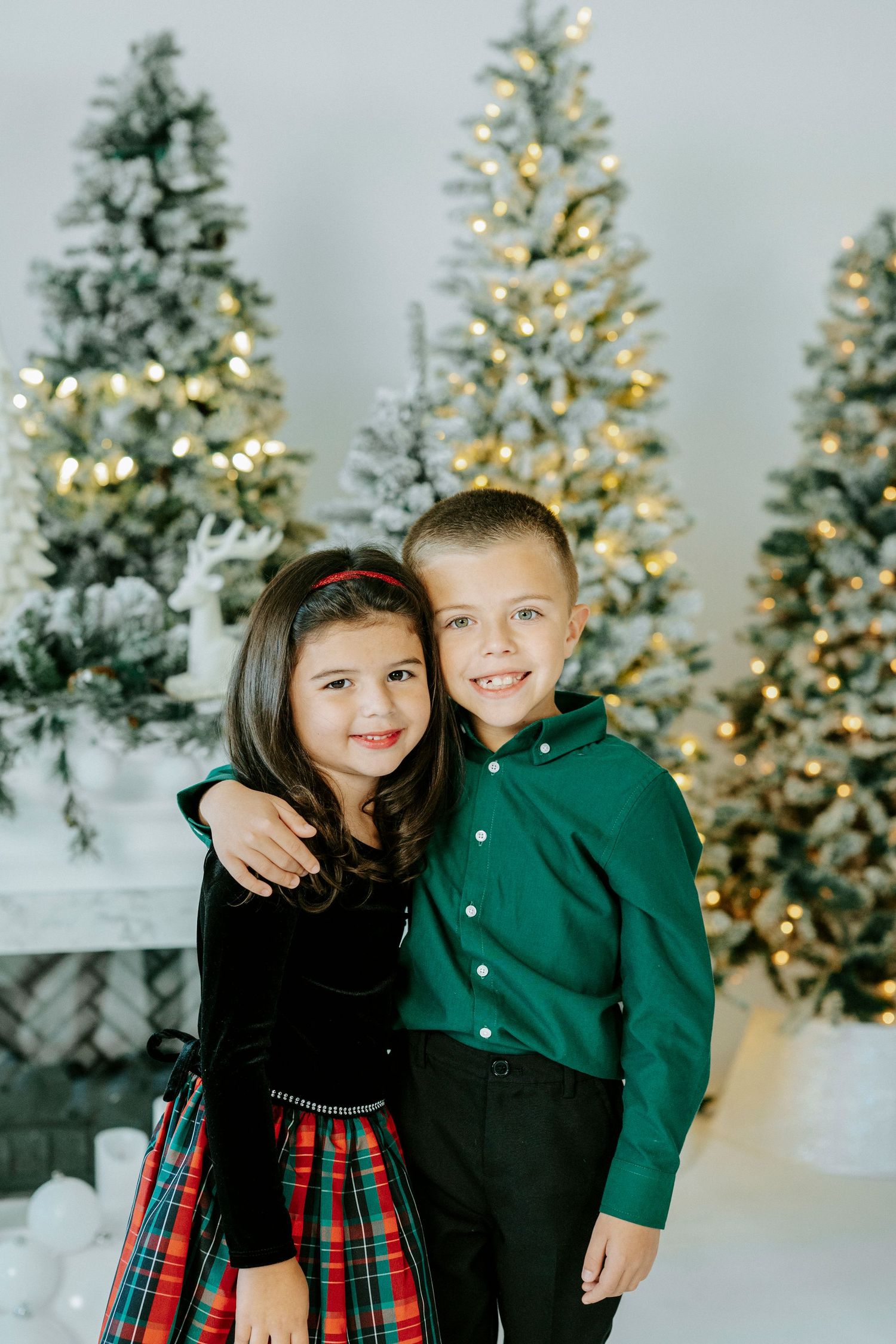 Two children in festive holiday outfits smile together in front of illuminated Christmas trees and a white fireplace.