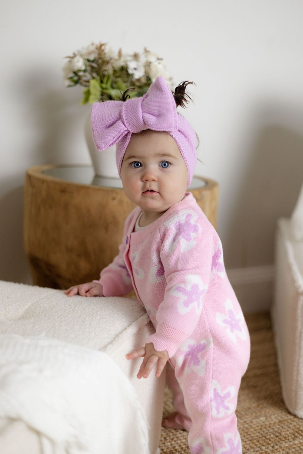 Adorable baby girl in pink floral romper and purple bow headband standing by a white couch.