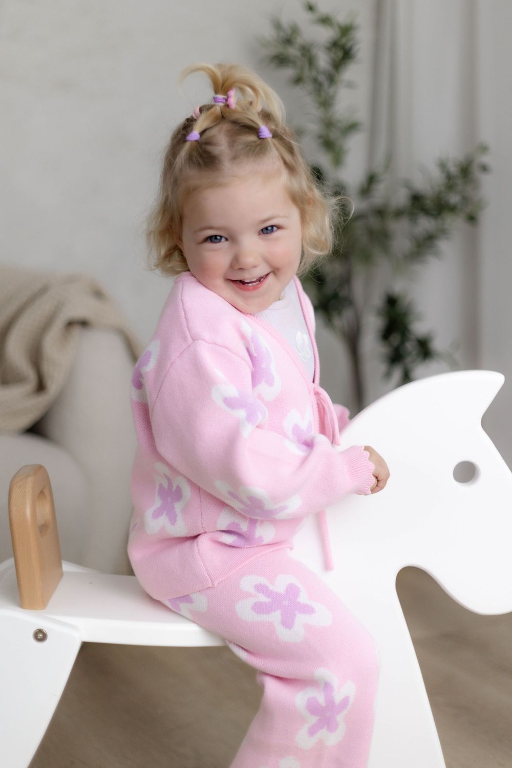 Smiling toddler girl in pink floral pajamas sitting on a white wooden rocking horse in a bright room.