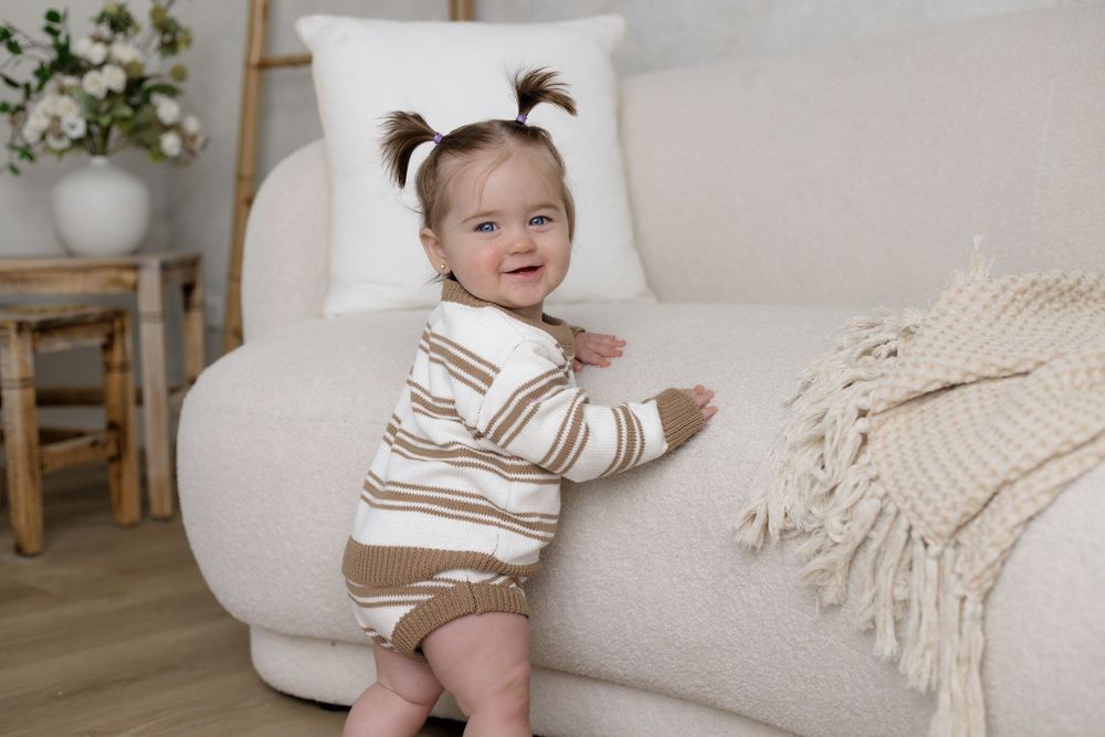 Cute baby girl in striped romper standing against a white boucle sofa in a bright, cozy living room.