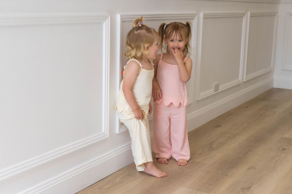 Two young girls in pastel pajamas whispering secrets while standing barefoot against a white wainscoting wall.