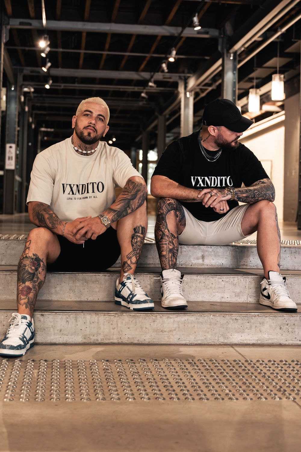 Two tattooed men in VXNDITO branded clothing sit on industrial steps.