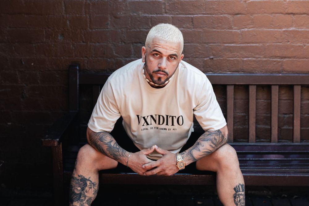 A tattooed man in a VXNDITO t-shirt sits on a wooden bench against a brick wall.
