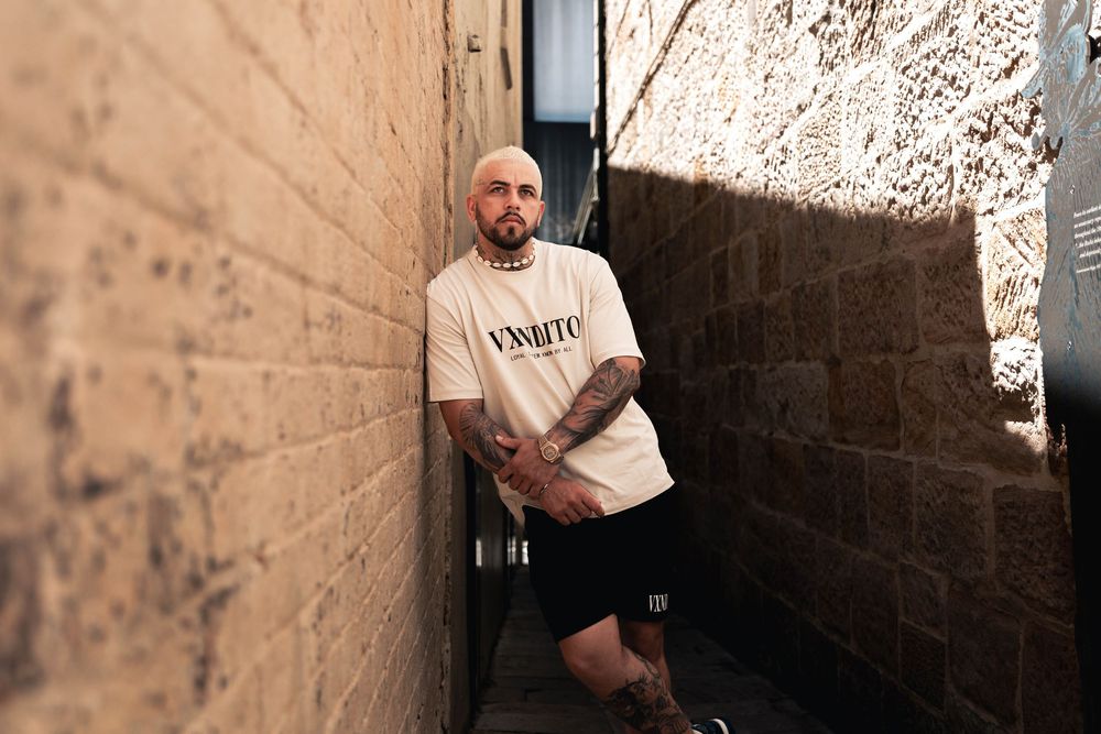 A man in VXNDITO apparel poses in an urban alleyway with textured walls.