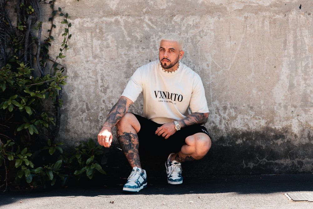 A streetwear model in VXNDITO clothing crouches against a concrete wall outdoors.