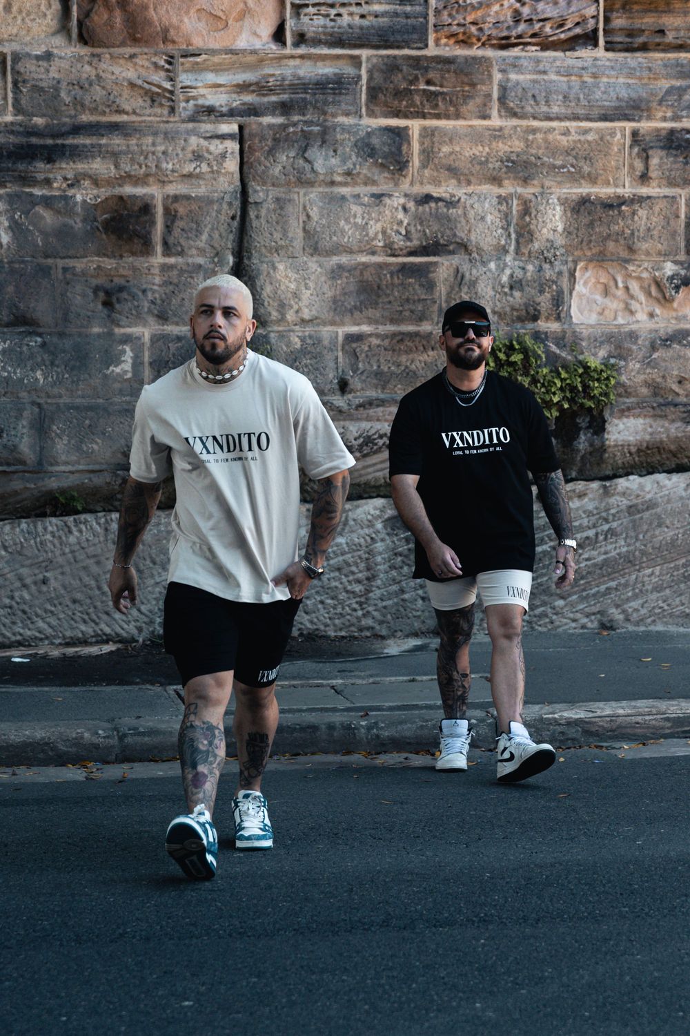 Two people in contrasting black and white streetwear walking along stone wall.
