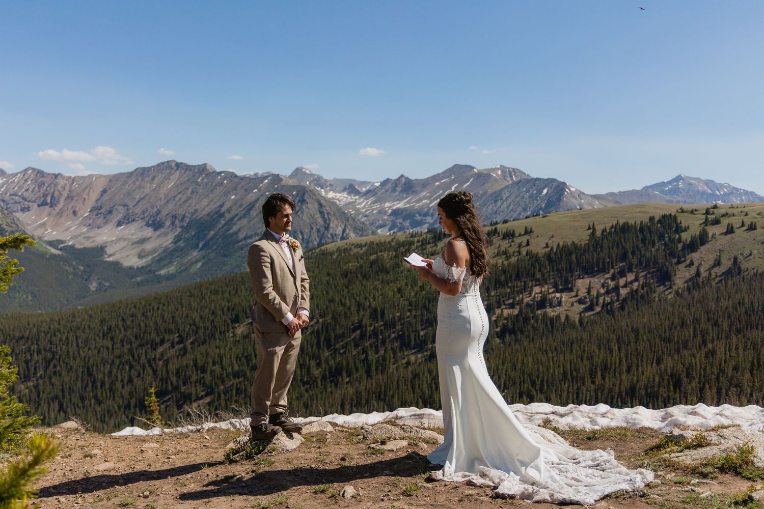 A couple in wedding attire stands on a scenic mountain overlook with dramatic peaks and valleys in the background.