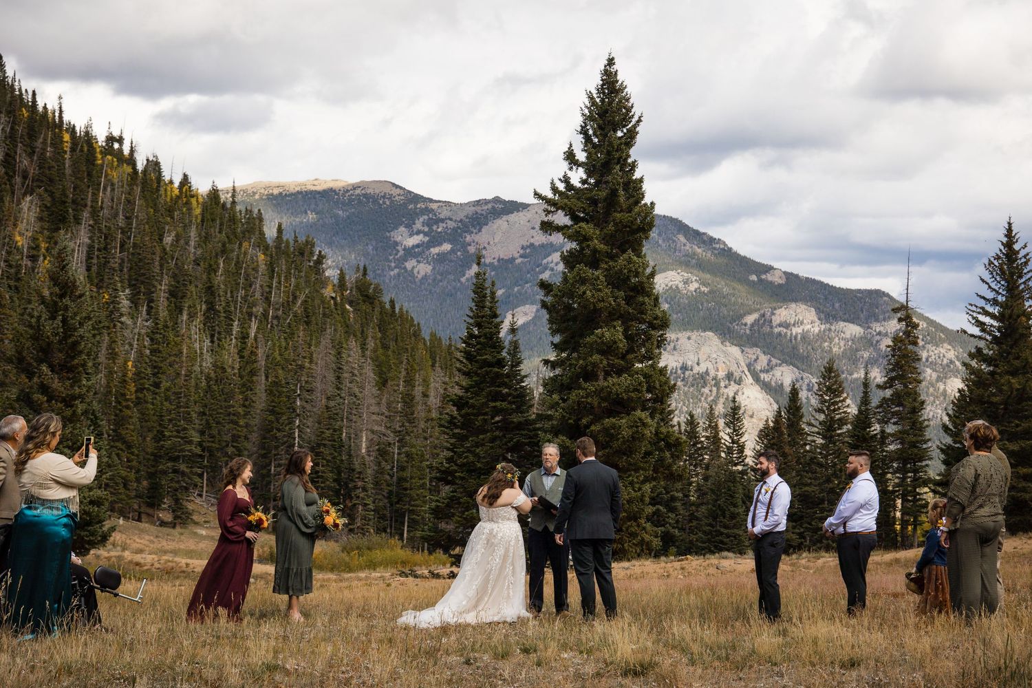 A small wedding ceremony takes place in a scenic mountain meadow surrounded by evergreen trees and snow-capped peaks.