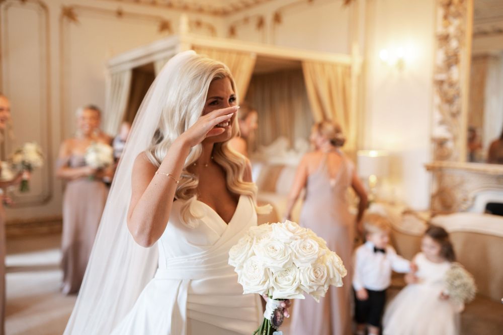A bride in an elegant white wedding dress walks down an ornate hallway with decorative moldings and chandeliers.