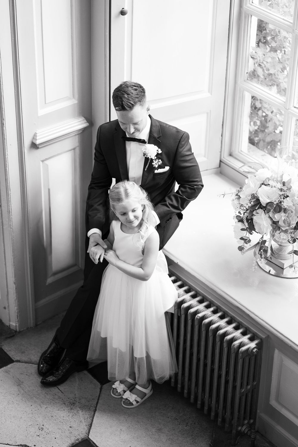 A tender black and white wedding moment captured by a window with a radiator and floral arrangement in view.