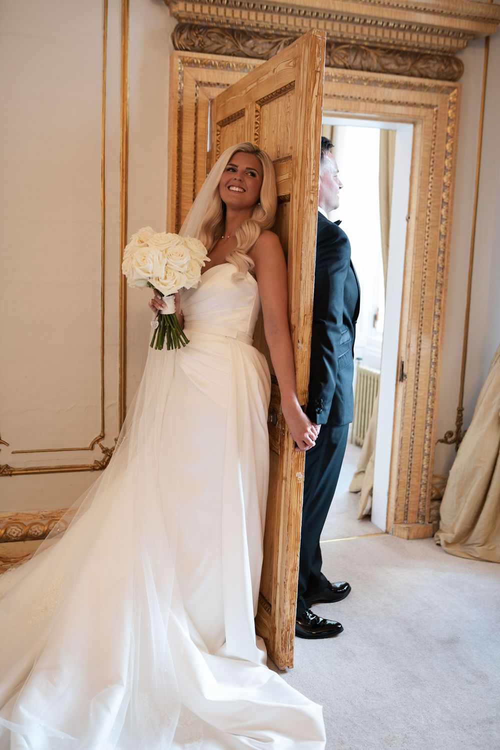 A bride in a white strapless wedding gown holds a bouquet of white flowers while standing in an elegant room with ornate doors.